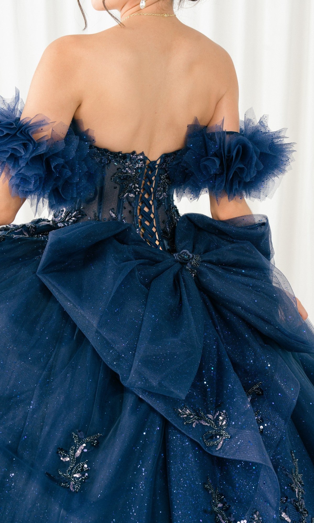 Person in the Quinceanera Dress 8380J By Cinderella Couture, a navy blue ball gown featuring an off-shoulder corset bodice, tulle ruffles, a large bow, and glittery floral embellishments.